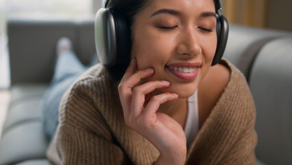 Hispanic latin woman relaxed girl Caucasian lady happy female lying on couch home apartment listen music headphones enjoy sound eyes closed sing song listening audio in earphones relax leisure smiling