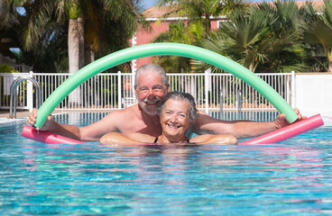 Cheerful happy senior couple having fun in outdoors swimming pool doing exercise with swim noodles....