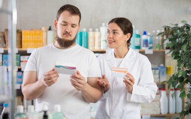 Obraz premium Young man buyer choosing box of pills with help of young woman pharmacist in pharmacy