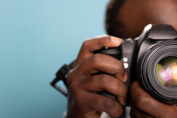 Extreme closeup of black man holding DSLR camera in studio with blue backdrop. Male photographer demonstrates camera handling techniques with selective focus on lens, emphasizing the intricate details