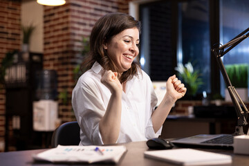Professional woman celebrates career milestone at desk, dancing in her chair with arms raised,...