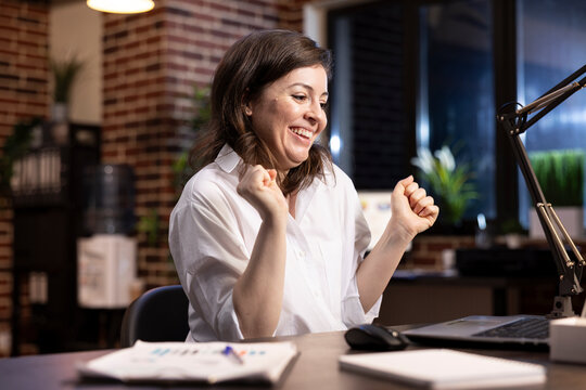 Professional woman celebrates career milestone at desk, dancing in her chair with arms raised, smiling as she looks at her laptop. Excited female entrepreneur feeling triumphant after an achievement.