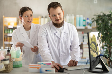Fototapeta premium Qualified male and female pharmacists work at a computer, renew stocks in the warehouse, enter new arrivals of medicines and pills. Pharmacists working behind the counter in a pharmacy