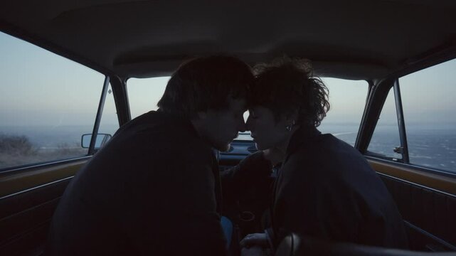 Romantic couple leaning in for tender forehead touch and nose rubs inside retro car parked near frozen lake during evening date. View from behind
