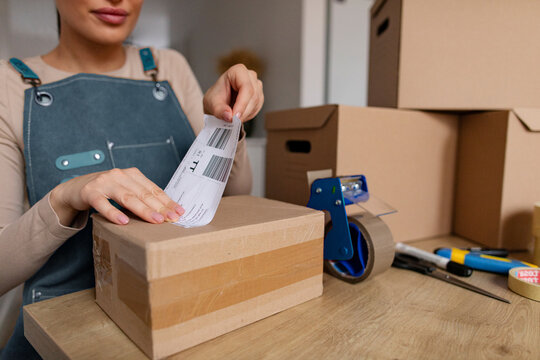 Young female online business owner putting label tag on parcel, preparing for delivery, working at table with cardboard boxes nearby