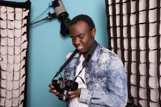 Professional male photographer holding camera and reviewing his pictures during a photoshoot session in studio. Young black cameraman standing and happily looking at images on his dslr screen.