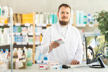 Obraz premium Young male pharmacist working at computer offering box of pills in pharmacy