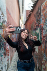 Young woman taking selfie with smartphone in graffiti alley, making peace sign
