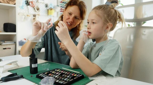 Young girl learns resin jewelry making at home with mother. Creative activity develops patience, fine motor skills, and artistic thinking, turning simple crafting project into educational experience.