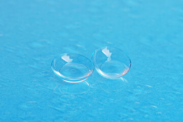 Pair of contact lenses and water drops on light blue mirror surface, closeup