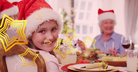 Image of stars falling over happy caucasian family wearing santa hats and having dinner