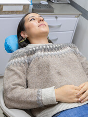 A young woman lies back in a dental or medical examination chair within a clinic setting, looking upwards with a calm, neutral expression