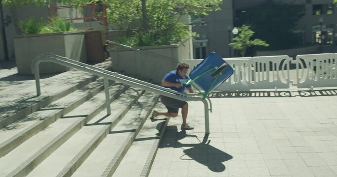 Funny shot of man trying to skid wheel barrow on rail like a skateboard then falling to ground - steady cam