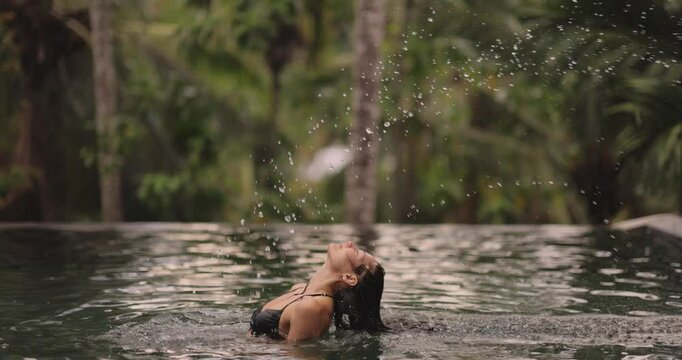 A stylish woman in a black bikini flips her wet hair in a tranquil tropical pool surrounded by lush greenery. Perfect for summer, travel, and luxury vacation concepts in exotic settings.