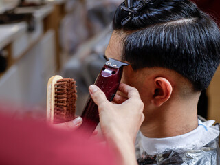 Asian man getting a precise undercut haircut at a barbershop. Barber uses red clipper to shape...
