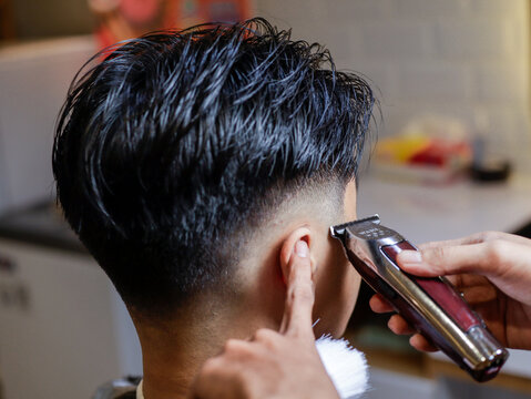 Asian male getting a sharp undercut at barbershop, hair clipper trimming around ear with barber's guidance. Clean fade hairstyle, dark textured hair, indoor lighting and salon background