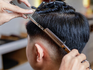 Asian male client in barbershop receiving haircut with scissors and comb. Hair sectioned with...