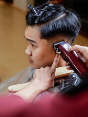 Asian man getting a haircut at a barbershop. Barber uses red clipper and wooden brush while...
