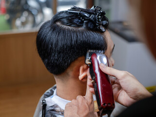 Asian man getting a haircut at a barbershop. Barber uses red clipper and wooden brush while pointing to fade line. Top hair held with black clips. Background shows mirror and warm wood decor