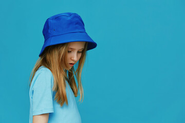 A young girl wearing a blue bucket hat and a matching t shirt, looking down against a solid blue background, embodying a calm and thoughtful mood © SHOTPRIME STUDIO