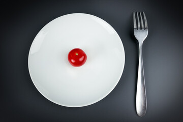 Small red tomato on a white plate, fork, gray background, close-up. The concept of dietary healthy eating, weight loss and vegetarianism.