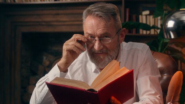 Satisfied old Caucasian man holding book textbook reading reader literature looking at camera smiling male home library pensioner grandfather elderly senior education study businessman business indoor