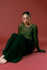 Fashion asian woman in black pants and sheer top sitting on red background with legs spread apart...