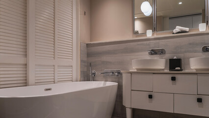 An oval bathtub in the corner. Faucets, shower on the tiled wall. There are wash basins, soap, a box of napkins on the countertop of the chest of drawers.The towels are rolled up.The blinds are closed