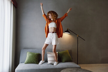 Joyful young woman in casual summer outfit celebrating at home, wearing white shorts and an orange...
