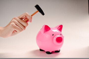 A human hand holding a small hammer raised above a glossy pink piggy bank on a cream background,...