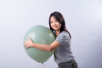 Fototapeta premium Asian girl with long black hair smiles while hugging a large round green balloon, wearing a striped blouse and dark skirt, standing sideways indoors against a clean white studio background