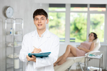 Friendly young male medical person writing at folder standing in treatment room of medical center.