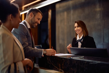 Happy businessman signing documents while checking in at hotel.