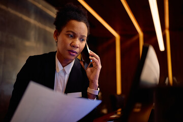 Black female hotel manager talking on cell phone at reception desk.