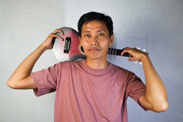 A handsome Asian young man in a t-shirt is holding a ukulele. The image shows a half-body view...