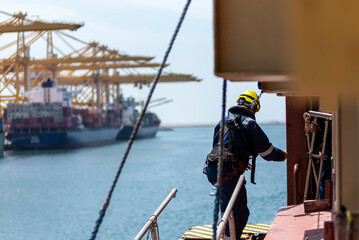 Fototapeta premium A deckhand prepares the pilot ladder as the ship readies for port entry. Secured in a safety harness, he carefully ensures the setup is ready, focusing on the task in a high-risk environment. 