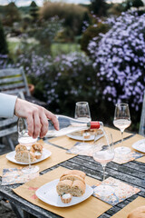 Hand pours rose wine into a glass on a rustic outdoor table set with bread and tarts in a floral garden. Culinary storytelling, outdoor brand shoot, rustic lifestyle branding, hospitality visuals