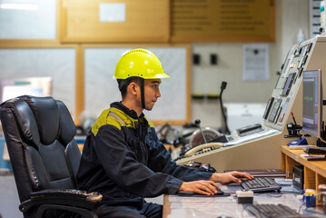 The marine engineer officer sits in the control room of a merchant vessel, focused on recording key operational parameters in the engine logbook, surrounded by monitors displaying system data.