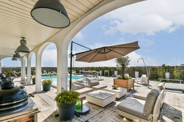 A stylish patio featuring cozy seating, elegant umbrellas, and a refreshing pool, surrounded by lush greenery and bright blue sky.