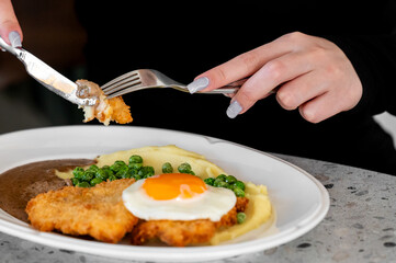 A close-up shot of a hand holding a fork and knife, preparing to eat a plate of fried chicken, mashed potatoes, green peas, and a sunny-side-up egg.
