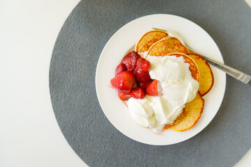 White round plate with pancakes, ice cream and red strawberries and a fork on the side on top of a round grey felt coaster on a white table, flat lay, right side