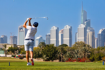 Golfer teeing off with city skyline backdrop, Male golfer swinging driver on a lush green golf course with a modern urban.