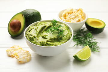 Tasty avocado dip in bowl, dill, lime and puffed rice cakes on white wooden table, closeup