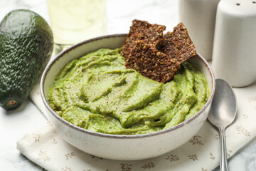 Tasty avocado dip in bowl, crispbreads and fruit on white marble table, closeup