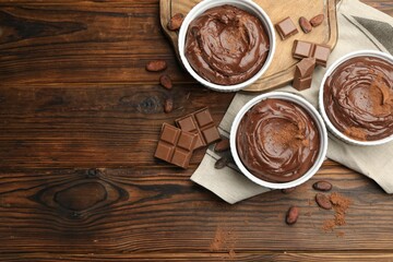 Tasty pudding in bowls, pieces of chocolate and cocoa beans on wooden table, flat lay. Space for...