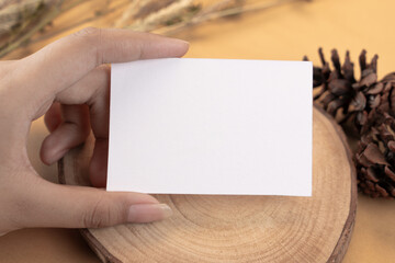 Obraz premium Hand holding white blank business card mockup with wooden decoration and dried flower on brown background