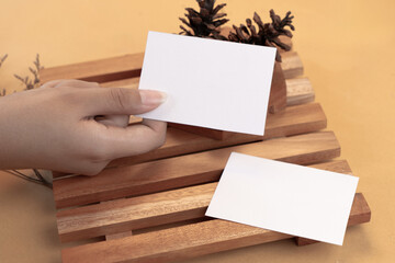 Obraz premium Hand holding white blank business card mockup with wooden decoration and dried flower on brown background