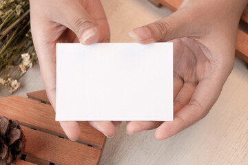 Obraz premium Hand holding white blank business card mockup with wooden decoration and dried flower on brown background