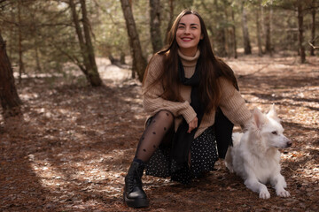 A woman in a cozy sweater and polka dot skirt kneels in the forest beside her white dog, smiling and gently petting it. The natural setting creates a serene and joyful moment.