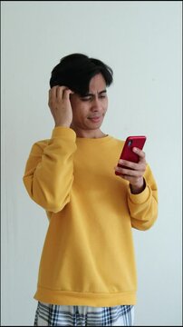 Confused young Indonesian Asian man scratching his head while looking at smartphone, expressing doubt or hesitation, isolated on white background, 4K resolution.